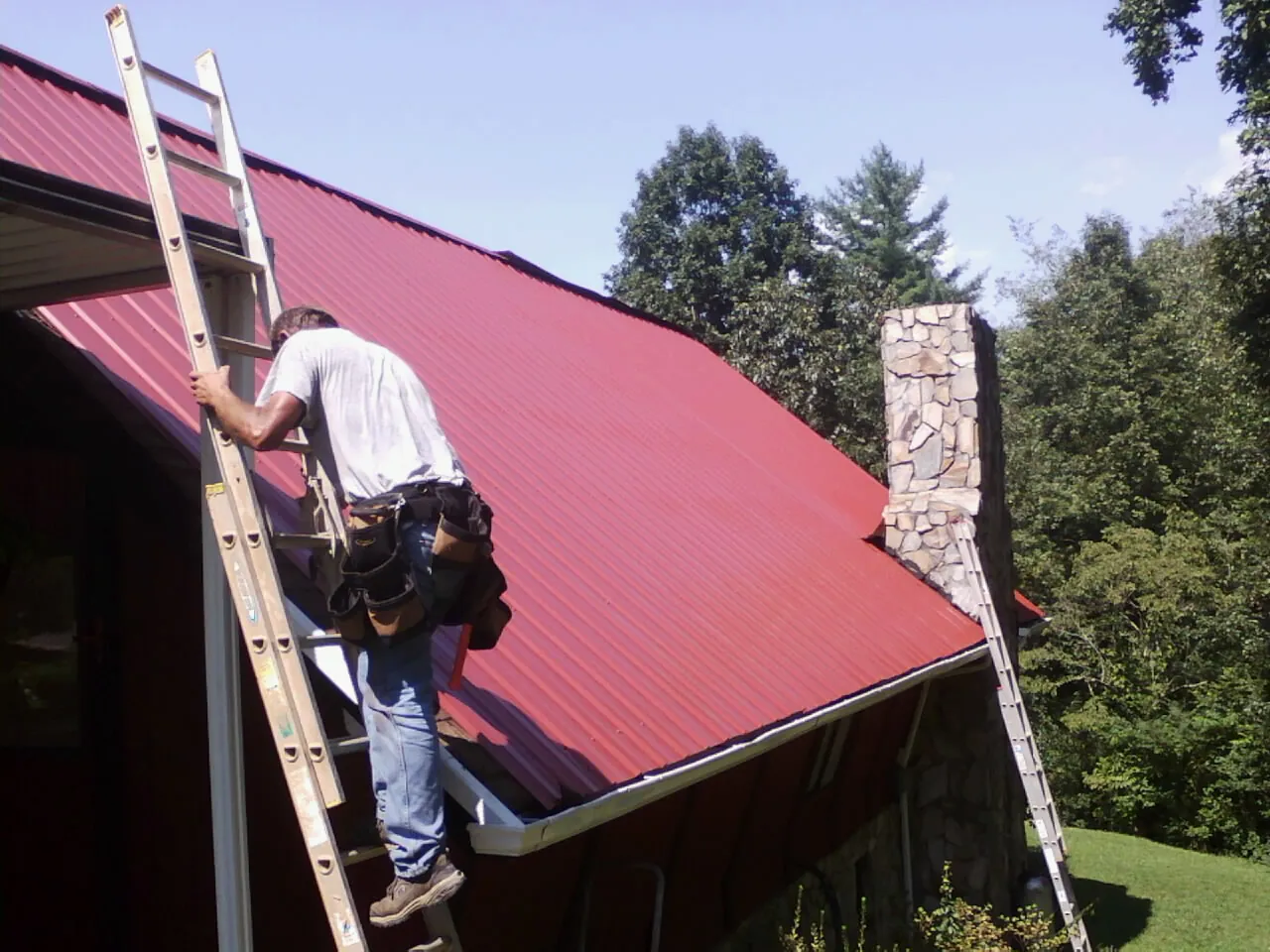 Roofer on a ladder beside a red metal roof and stone chimney — Soffit & Fascia Repair by Roofing Brothers in Cape Elizabeth